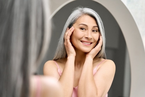 woman with gray hair looking at mirror touching face woman with gray hair looking at mirror touching face